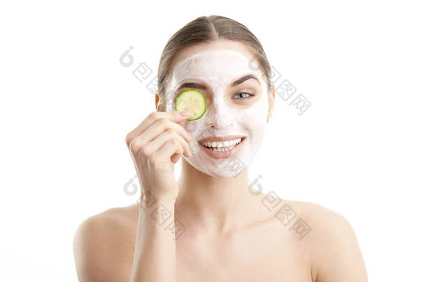 特写镜头, 一个微笑的美丽的年轻女子戴着口罩, 在她的眼睛面前拿着黄瓜片。在白色背景上被隔离.
