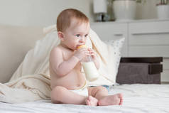 可爱的小男孩在尿布上坐在床上, 喝牛奶从瓶子的肖像