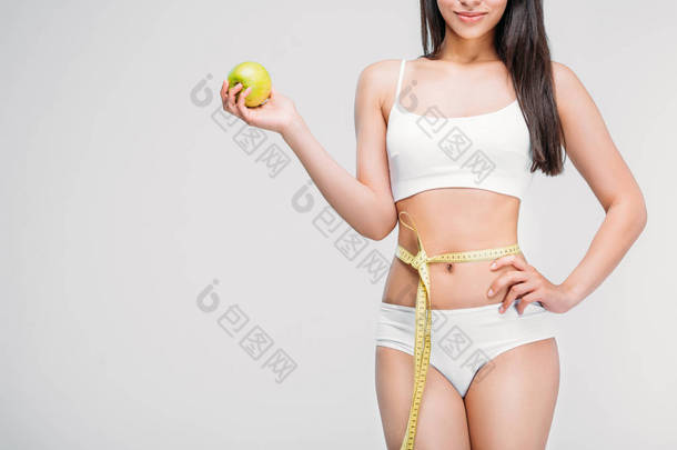 年轻苗条的女人在内衣与测量胶带在腰部举行苹果, 孤立的灰色, 饮食概念的裁剪视图