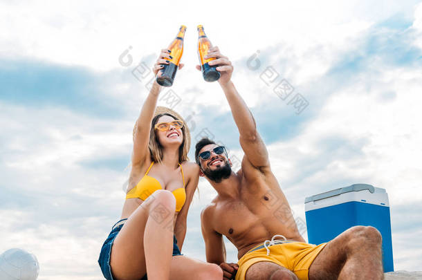 美丽的年轻夫妇的底部视图在乌云密布的天空前, 在沙滩上举起啤酒瓶