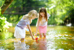 两个小妹妹在温暖晴朗的夏日里在河边玩纸船。孩子们在水里玩得很开心。幼儿暑期活动.
