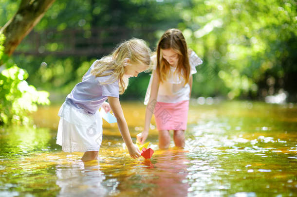 两个小妹妹在温暖晴朗的夏日里在河边玩纸船。孩子们在水里玩得很开心。幼儿暑期活动.