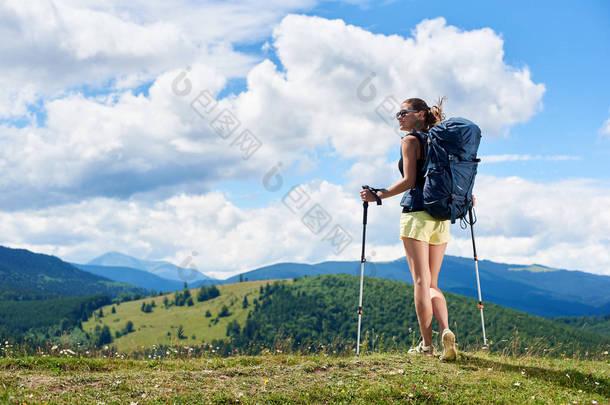 运动员微笑的女子旅游徒步登山小径, 走在草地上, 穿着背包和太阳镜, 使用徒步棍, 享受夏日阳光明媚的一天在山上。旅游理念