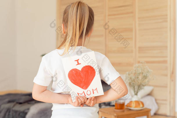 小女孩抱着贺卡妈妈在母亲节的背面与题词我爱妈站在卧室里 , 母亲日概念