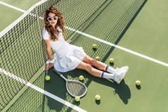 青年时尚网球运动员的高角度观看白色运动衫和太阳镜坐在网上与网球设备在网球场附近