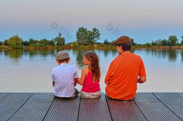 坐在码头上的孩子们。三名不同年龄的儿童-十几岁男孩, 小学生和学龄前女孩坐在一个木码头。<strong>夏日</strong>和童年的概念。孩子们在<strong>湖边</strong>的长凳上。假期晚上的黄金时刻.