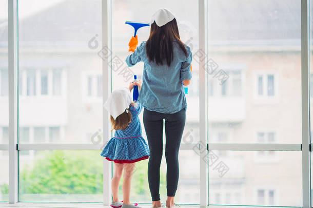 洗窗户的年轻母亲的背影和她的小女儿在室内帮助母亲