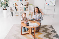 快乐的心理学家坐在小孩旁边的剪贴板上, 而她则用彩色铅笔画画, 看着相机