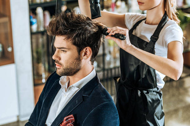 裁剪拍摄的年轻美发师梳理和干燥的头发英俊的客户在美容院