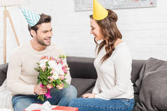 英俊的微笑的男人在党的帽子提出生日鲜花的女人在客厅