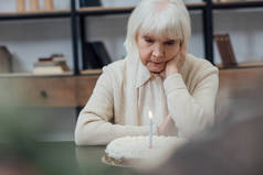 心烦意乱孤独的老年妇女坐在桌旁, 看着生日蛋糕与燃烧的蜡烛在家里