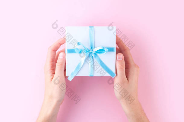 女性手拿着一个白色的小礼品盒包裹着蓝色的丝带在粉红色的背景。给予和接受来自亲人的礼物.