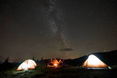 公司由五名快乐的游客用吉他旅行, 男人和女人坐在帐篷附近燃烧的篝火旁, 享受美丽的野营之夜