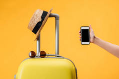 裁剪的妇女持有智能手机与空白屏幕靠近旅行袋, 草帽和太阳镜隔离在黄色