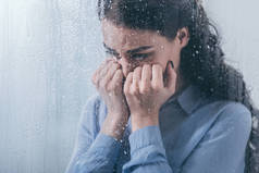郁闷的女人用双手捂住脸, 用雨滴在家里透过窗户哭泣