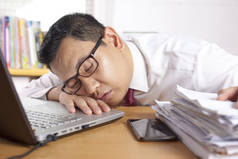 亚洲商人因过度劳累而疲惫不堪, 在办公室的笔记本电脑和纸制品前睡觉的肖像