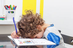 累了的小男孩坐在桌旁, 一边做作业, 一边在笔记本上写字。学校大量作业的概念