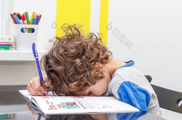 累了的小男孩坐在桌旁, 一边做<strong>作业</strong>, 一边在笔记本上写字。学校大量<strong>作业</strong>的概念