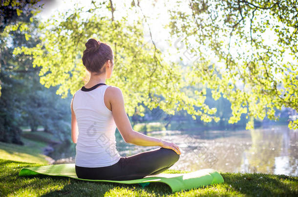 年轻女子<strong>打坐</strong>, 而坐在瑜伽毯在上午公园附近的湖, 在街头锻炼的概念