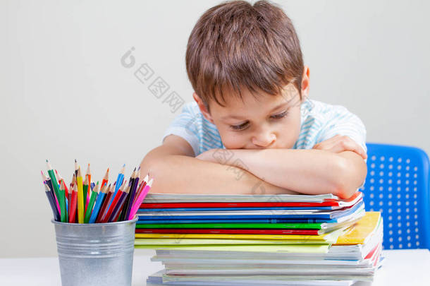 不开心的小学生坐在课桌上, 堆着书本和笔记本。