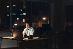 年轻的亚洲商人在一台笔记本电脑上工作, 而晚上坐在他的办公桌在黑暗的办公室与城市灯光背景