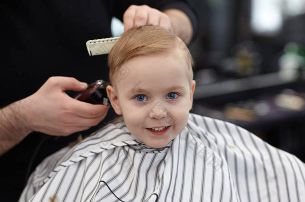 可爱的金发碧眼的微笑的男婴与蓝色的眼睛在理发店有理发由<strong>美发</strong>师。有工具的造型师之手。<strong>儿童</strong>时尚。室内, 黑暗的背景, 复制空间.