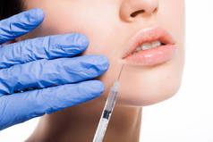在蓝色乳胶手套的美容师的裁剪视图举行注射器附近的年轻妇女的嘴唇隔离在白色