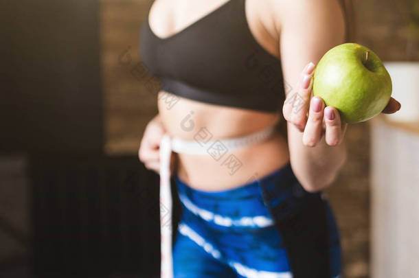一位穿着运动服装的美丽年轻女子测量测量的飞行腰围, 一手拿着一个苹果。运动、饮食和减肥