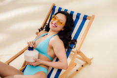 美丽的快乐女孩在比基尼和太阳镜与椰子鸡尾酒躺在甲板上的椅子上海滩