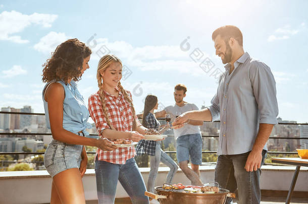美味的食物给最好的<strong>朋友</strong>。年轻快乐的男人站在屋顶上，把热烤肉放在他的<strong>女性朋友</strong>们的盘子里。烧烤的概念.