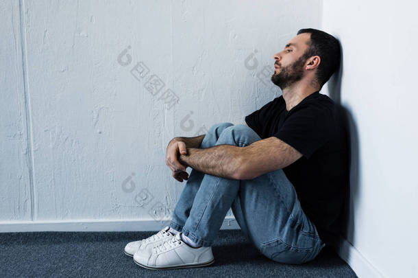 英俊的沮丧的人坐在地板上的角落，由白色的墙壁，看着远