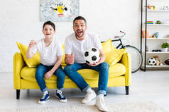 父亲和儿子欢呼，而观看体育比赛在沙发上与足球在家里