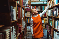 金发和美丽的女人在橙色礼服拿着书，看着在图书馆