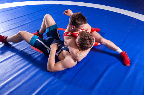 两名身着红色和蓝色制服的年轻男子<strong>摔</strong>跤手在体育馆的蓝色<strong>摔</strong>跤地毯上<strong>摔</strong>跤。拼杀.