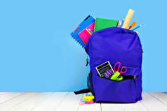 紫色背包里装满了蓝色背景的学校用品。回到学校的概念。复制空间.