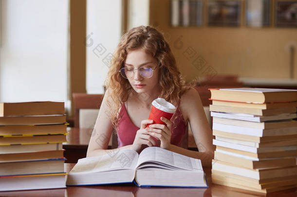 教育和学校理念。聪明的女学生手拿咖啡，戴着眼镜，穿着休闲装，神情专注，坐在大学图书馆的书桌上，拿着书堆看书.