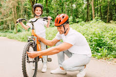 父亲在头盔检查自行车的车轮，而儿子站在头盔，看着远