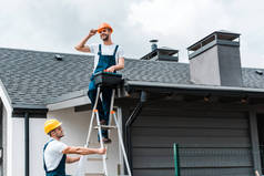 快乐的修理工坐在屋顶上，拿着工具箱靠近头盔的同事