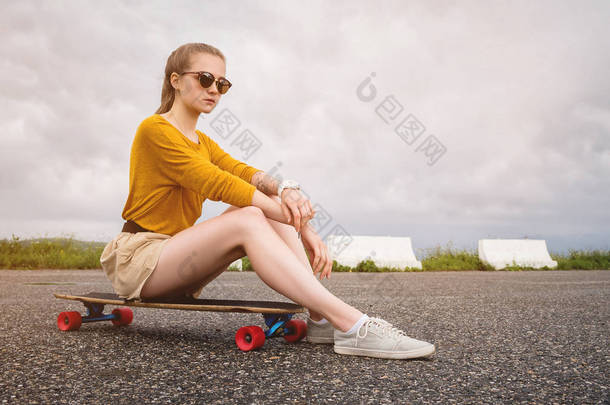 在灰色天空的背景中，戴着太阳镜和<strong>短裤</strong>的迷人年轻女孩坐在她的长板上。千禧一代与<strong>休闲</strong>理念