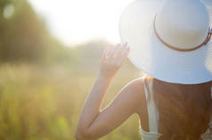 年轻的女孩在田野前戴着一顶明亮的大帽子，看着夕阳。背光，选择性对焦。戴帽子的年轻女子看着太阳。复制空间
