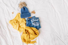 舒适的黄色毛衣，蓝色牛仔裤，围巾和帽子在床上的白床单。女士时尚的秋冬套装。时髦的服装拼贴画。平铺，顶视图.