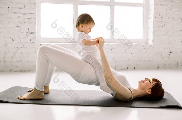 年轻快乐的妈妈锻炼身体，做<strong>臀</strong>部桥<strong>运动</strong>，穿着白色<strong>运动</strong>服，肚子上的小女孩，健康，产后瑜伽。 健康生活方式概念