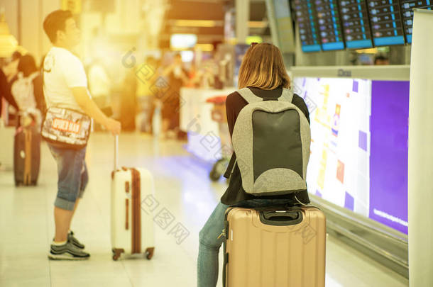 有选择地把重点放在女游客的背像上，带着模糊的男性乘客坐在行李箱上，看着背景下的航班时刻表监视器