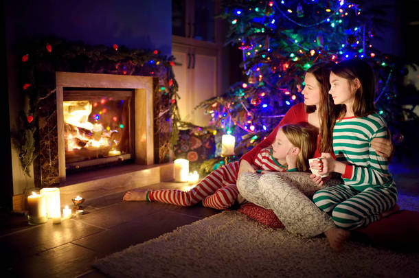平安夜，快乐的年轻妈妈和她的女儿们一起坐在一个温暖的黑暗客厅的壁炉边，度过了一个愉快的夜晚。 在家里庆祝圣诞节.