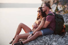 抱着一对背着背包的情侣坐在岩石山顶上，享受着沿江或湖景的美景。山区和海岸旅行、自由和积极的生活方式概念.