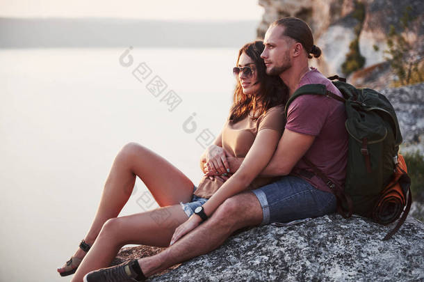 抱着一对背着<strong>背包</strong>的<strong>情侣</strong>坐在岩石山顶上，享受着沿江或湖景的美景。山区和海岸旅行、自由和积极的生活方式概念.