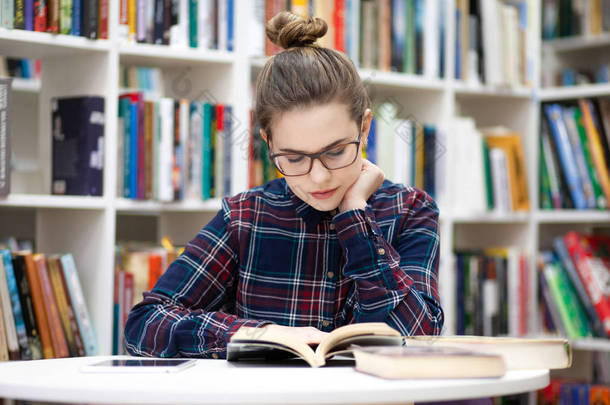 女孩坐在图书馆里。 穿着格子衬衫戴眼镜的年轻学生坐在公共图书馆看书。 女人喜欢看书