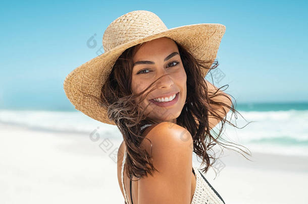 <strong>在</strong>背景为大海的<strong>海滩上</strong>,一个戴着草帽的美丽微笑的年轻女子的画像.漂亮<strong>女孩</strong>笑着看着海滨的相机。一个无忧无虑的晒黑了的女人走<strong>在</strong>沙滩<strong>上</strong>笑着.