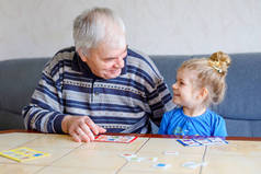 漂亮的蹒跚学步的女孩和爷爷一起在家里玩图片彩票桌卡游戏。可爱的孩子和老年人在一起玩得很开心.在室内快乐的家庭