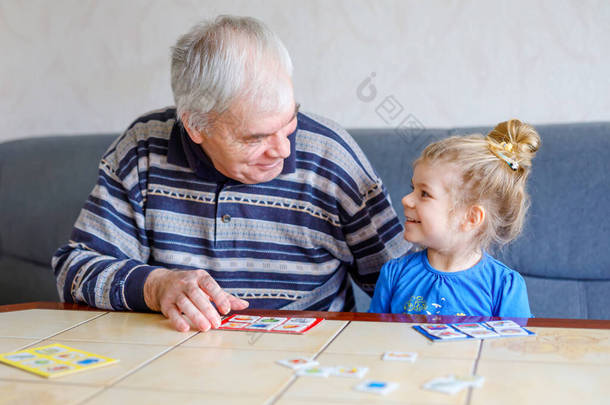 漂亮的蹒跚学步的女孩和爷爷一起在家里玩图片彩票<strong>桌卡</strong>游戏。可爱的孩子和老年人在一起玩得很开心.在室内快乐的家庭
